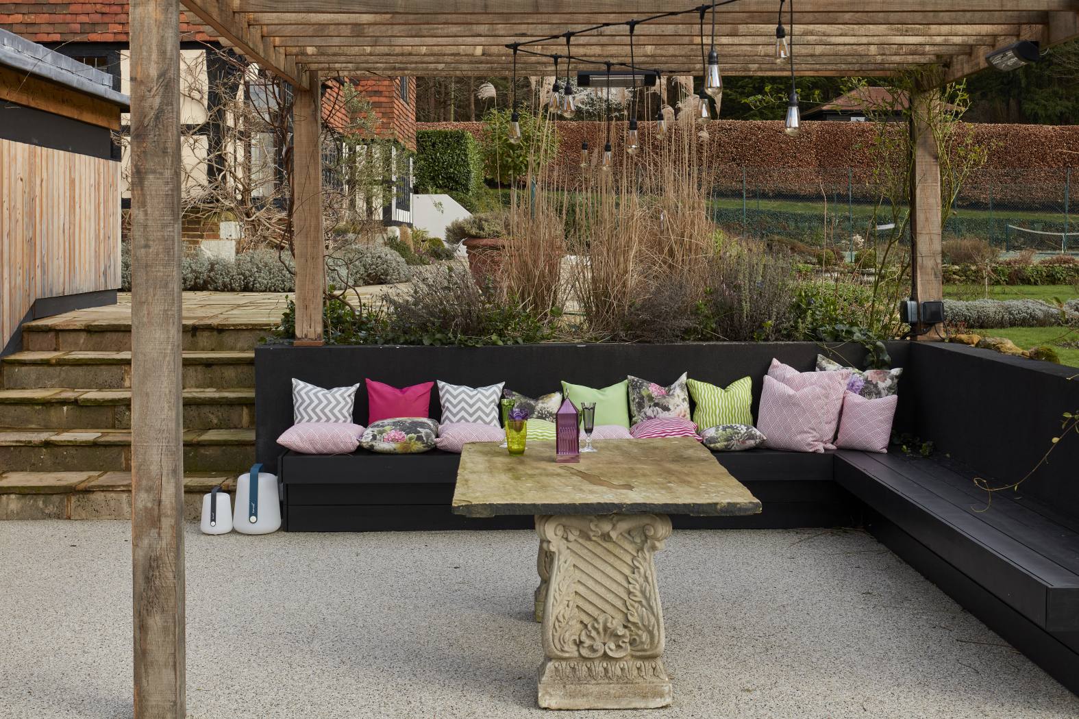 Garden seating area under a wooden pergola with colourful cushions and an antique stone table.