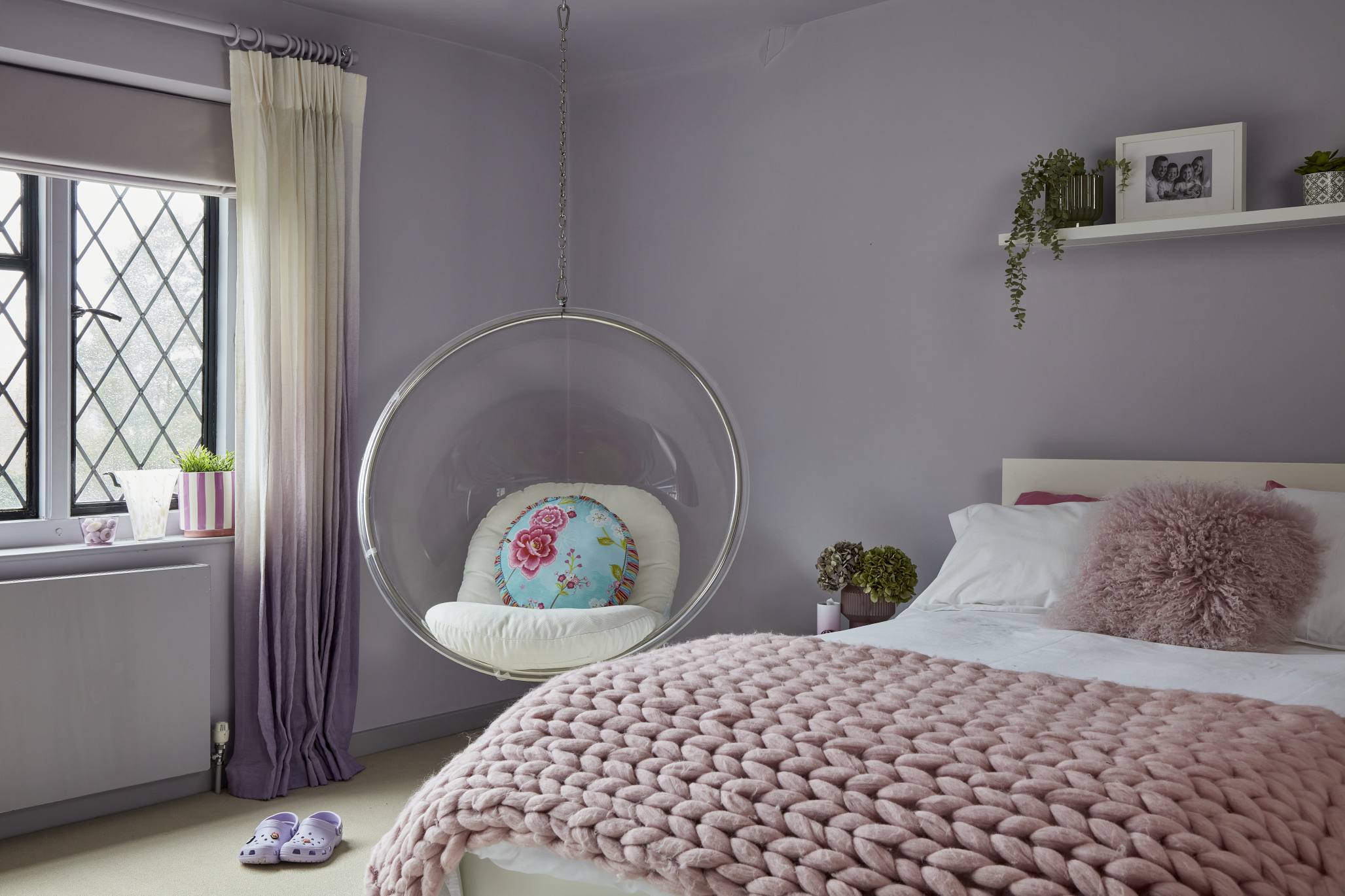 A lavender-painted girl's bedroom with a hanging bubble chair, a white bed with pink accents, and ombre curtains.