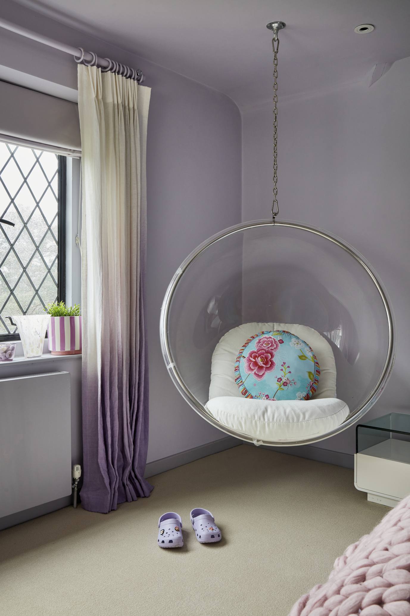 A neatly styled girl's bedroom with a hanging bubble chair, white bedside table, and plush pink and white bedding.