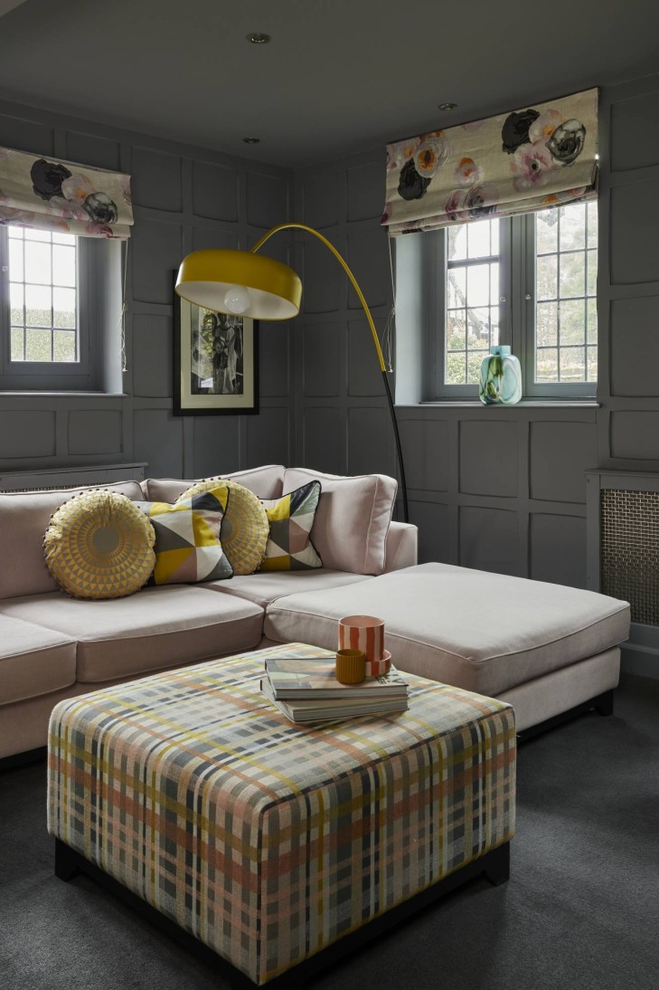 Luxury home library with soft grey panelled walls, pink chaise sofa, plaid ottoman, and a bold yellow arc floor lamp.