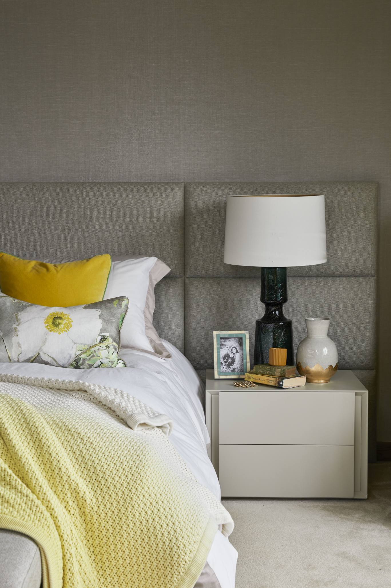 Close-up of a bedside table with soft neutral linens, yellow and floral pillows, and ambient lighting.